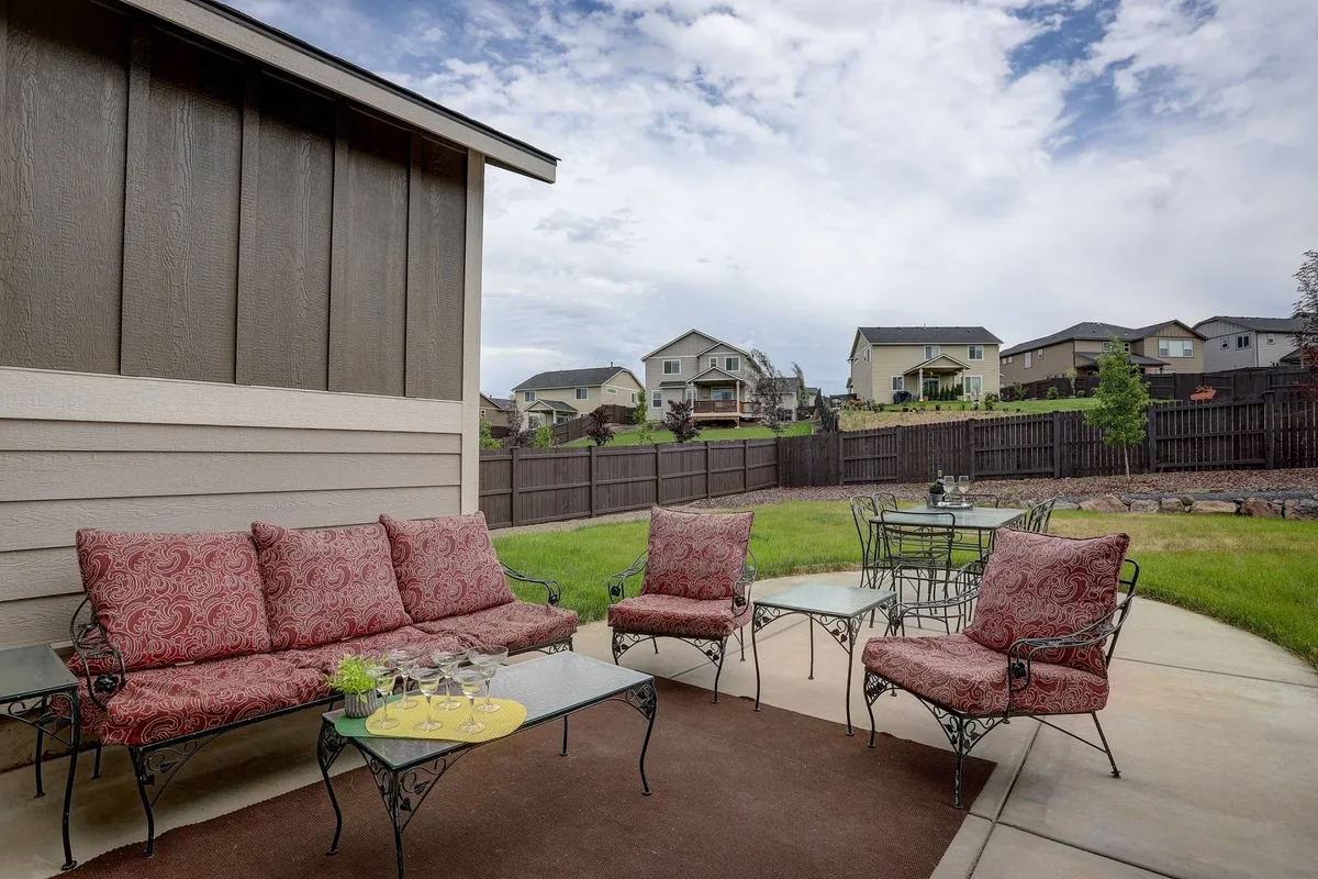 Red cushioned seating and a glass table are arranged on the patio, overlooking a green lawn and enclosed by a backyard fence. The scene appears welcoming and is perfectly positioned for enjoying sunset views or watching children play in the yard.