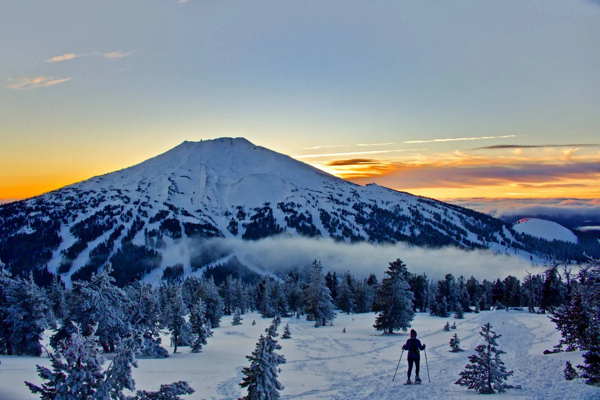 A panoramic outdoor scene featuring a snow-covered mountain bathed in the warm glow of sunset. A lone person skis in the foreground, with the sky painted in serene hues of orange and pink, evoking a sense of adventure and tranquility.