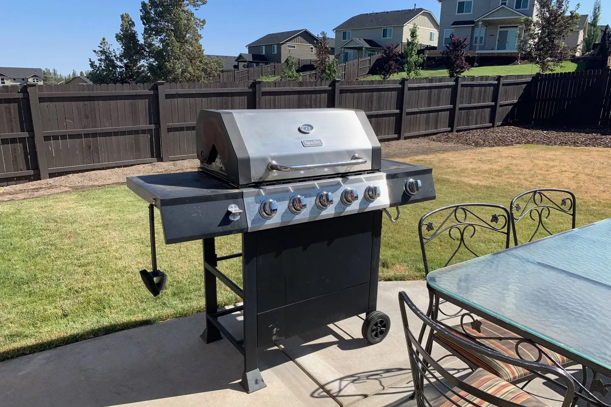A close-up of a grill and outdoor dining set on a patio, set against a backdrop of a fenced yard. The scene conveys a relaxed, social atmosphere, perfect for gatherings, barbecues, or al fresco dinners under the open sky.