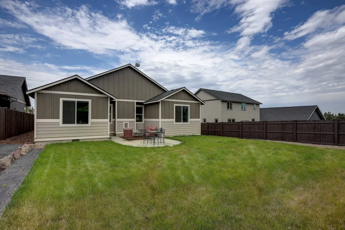 A wide view of the backyard showing a lush lawn, inviting patio seating, and the exterior facade of the house. The scene is bathed in the gentle light of either sunset or sunrise, evoking a feeling of tranquility and openness. The patio area is furnished and ready for relaxation or dining, while the spacious lawn offers ample space for play or leisure.