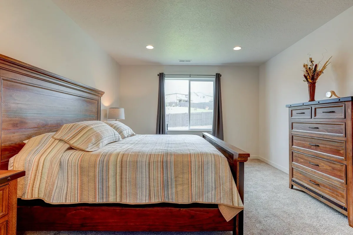 A peaceful bedroom featuring a cozy bed with plush bedding, a dresser, and a large window that lets in soft natural light. The serene color palette and minimal décor create a restful atmosphere ideal for relaxation.
