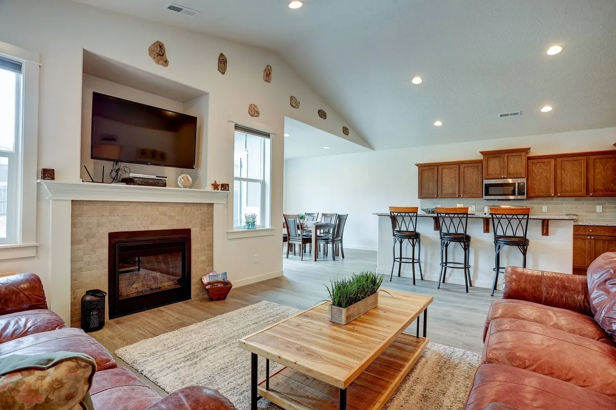 The image features a warm, inviting open-concept living space with a crackling fireplace, plush sofa, elegant dining area, and a modern kitchen with bar stools. The overall mood is cozy and welcoming, ideal for relaxation and gatherings.