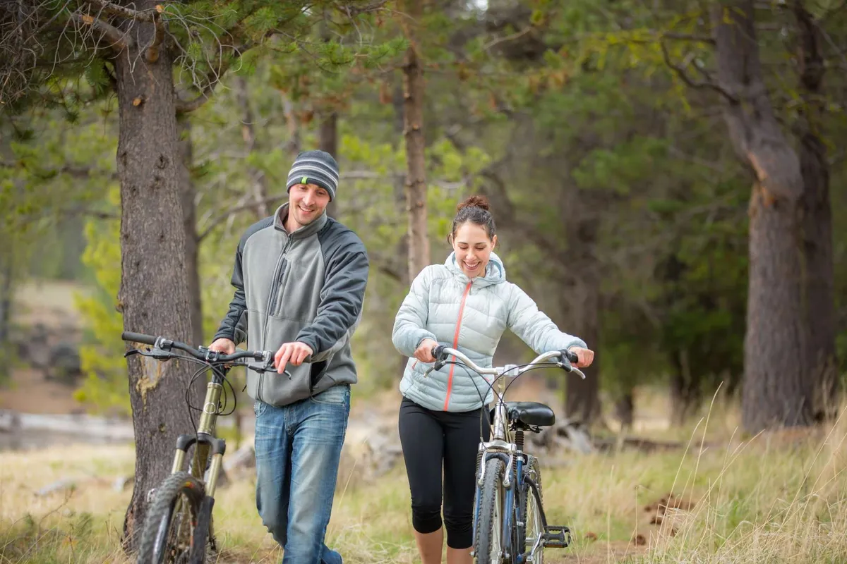 Two cyclists walk their bicycles along a scenic, tree-lined path with grassy fields on either side, evoking a sense of exploration and outdoor adventure.