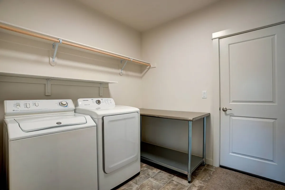 This interior image centers on a practical laundry room setup, featuring a full-size washing machine, drying machine, a shelf for storage, and a metal table for sorting and folding clothes. The scene conveys tidiness, convenience, and ease of use.