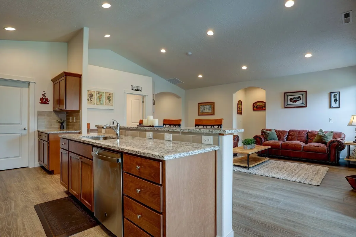This image features a spacious open-concept area highlighting the kitchen island with granite countertops, seamlessly flowing into the living room furnished with comfortable seating. The room is bathed in natural light, emphasizing the unified, sociable layout ideal for group gatherings and shared activities.