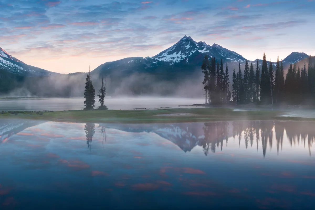 A tranquil vista featuring a glassy lake reflecting the surrounding mountains and a pastel sky. The scene is framed by trees, emphasizing peace, connection to nature, and the possibility of outdoor exploration.
